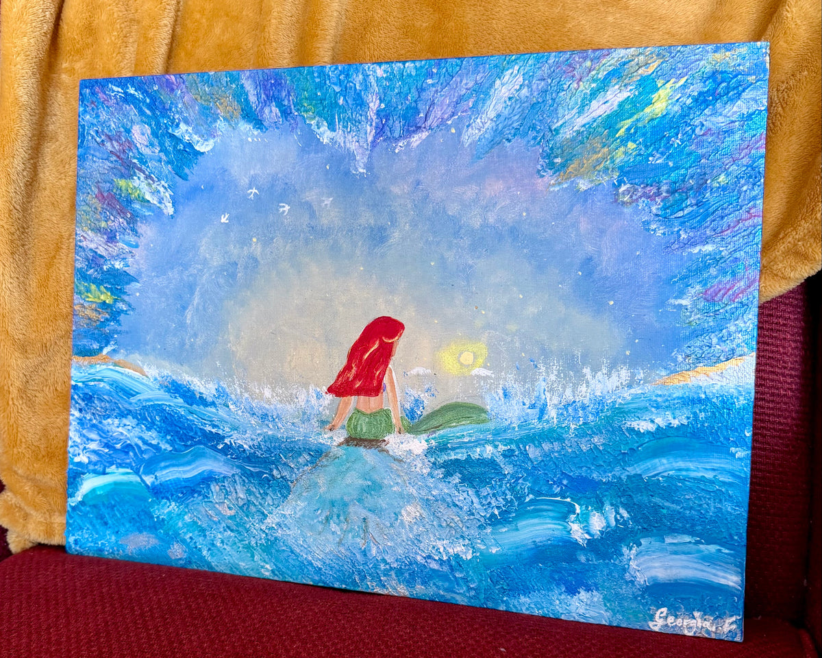 A colorful acrylic painting of a mermaid with red hair standing on a surfboard in a blue ocean, displayed on a red upholstered chair with a yellow blanket in the background.
