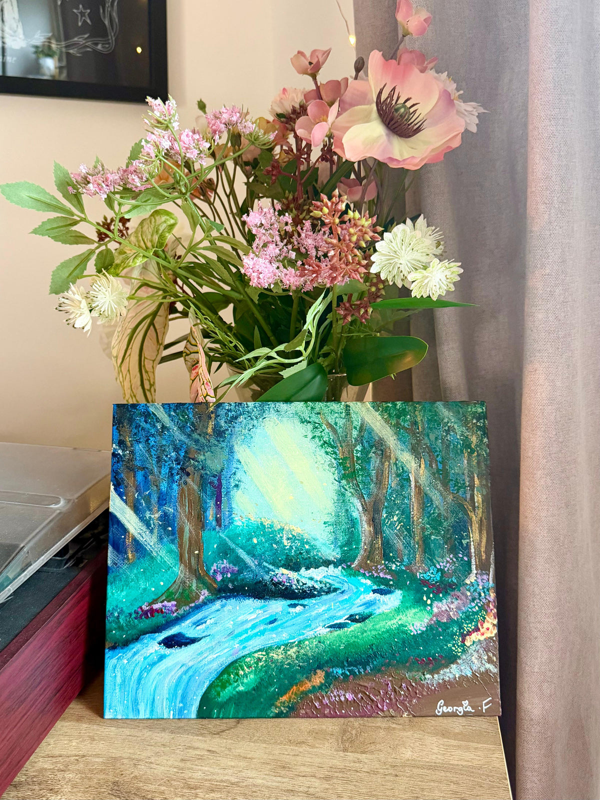 A colorful acrylic painting of a forest stream scene with green and blue foliage, displayed on a wooden surface next to a vase of pink and white flowers.