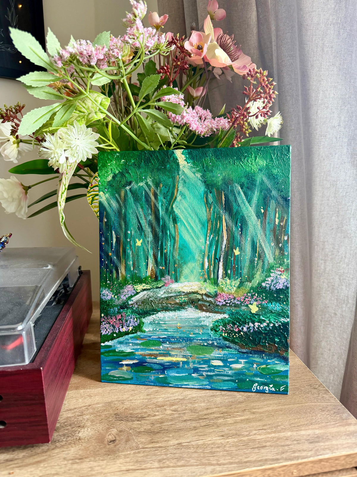 A small vertical canvas painting of a glowing green forest stream, styled in a vase with pink and white flowers on a wooden table next to a record player, with a gray curtain in the background.