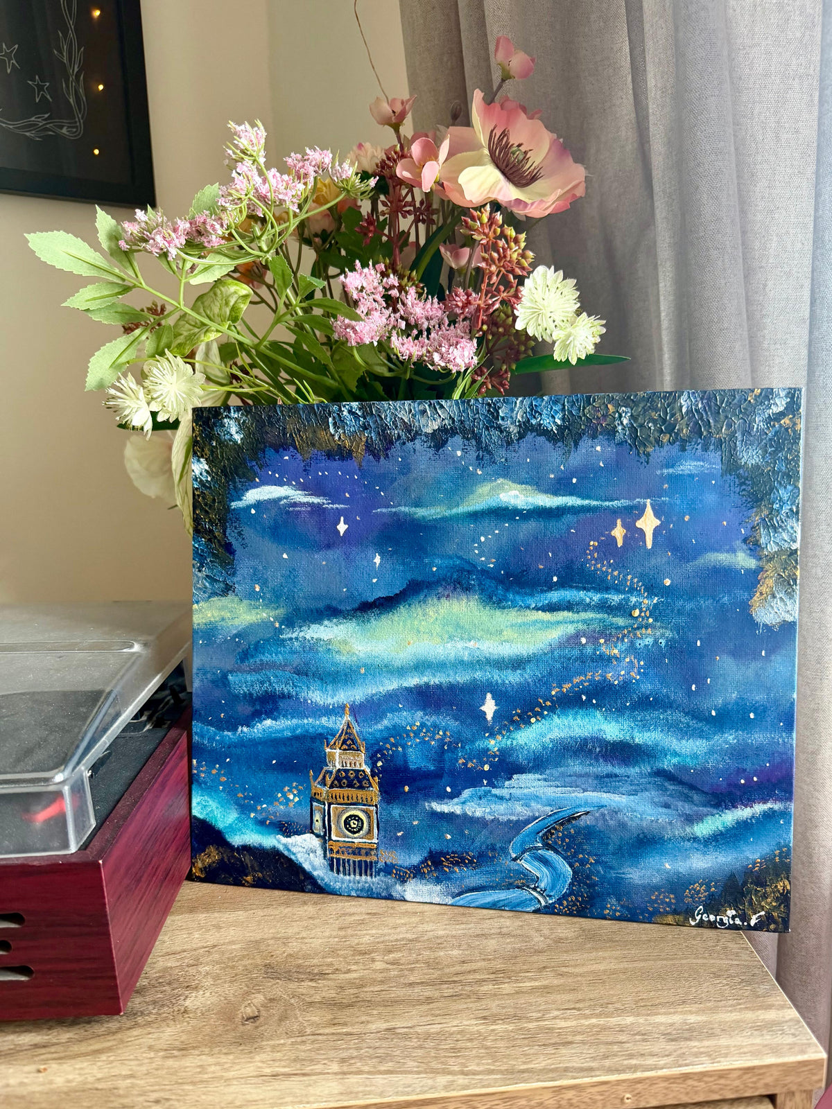 A square canvas painting depicting a whimsical blue night landscape with a tower and stars, styled on a wooden table next to a vase of flowers and a record player.