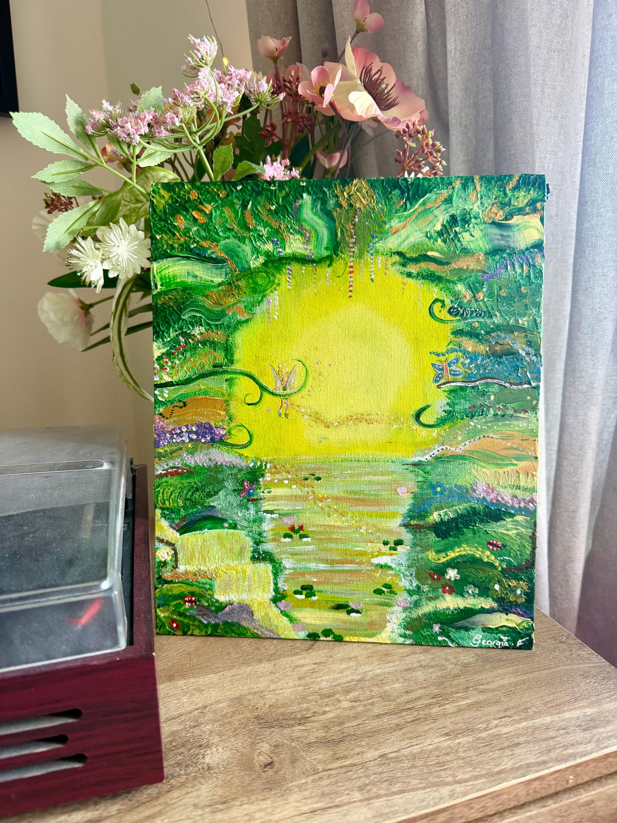 A vertical acrylic painting of a lush green landscape with a bright yellow sun and a winding river, displayed on a wooden table next to a vase of flowers.