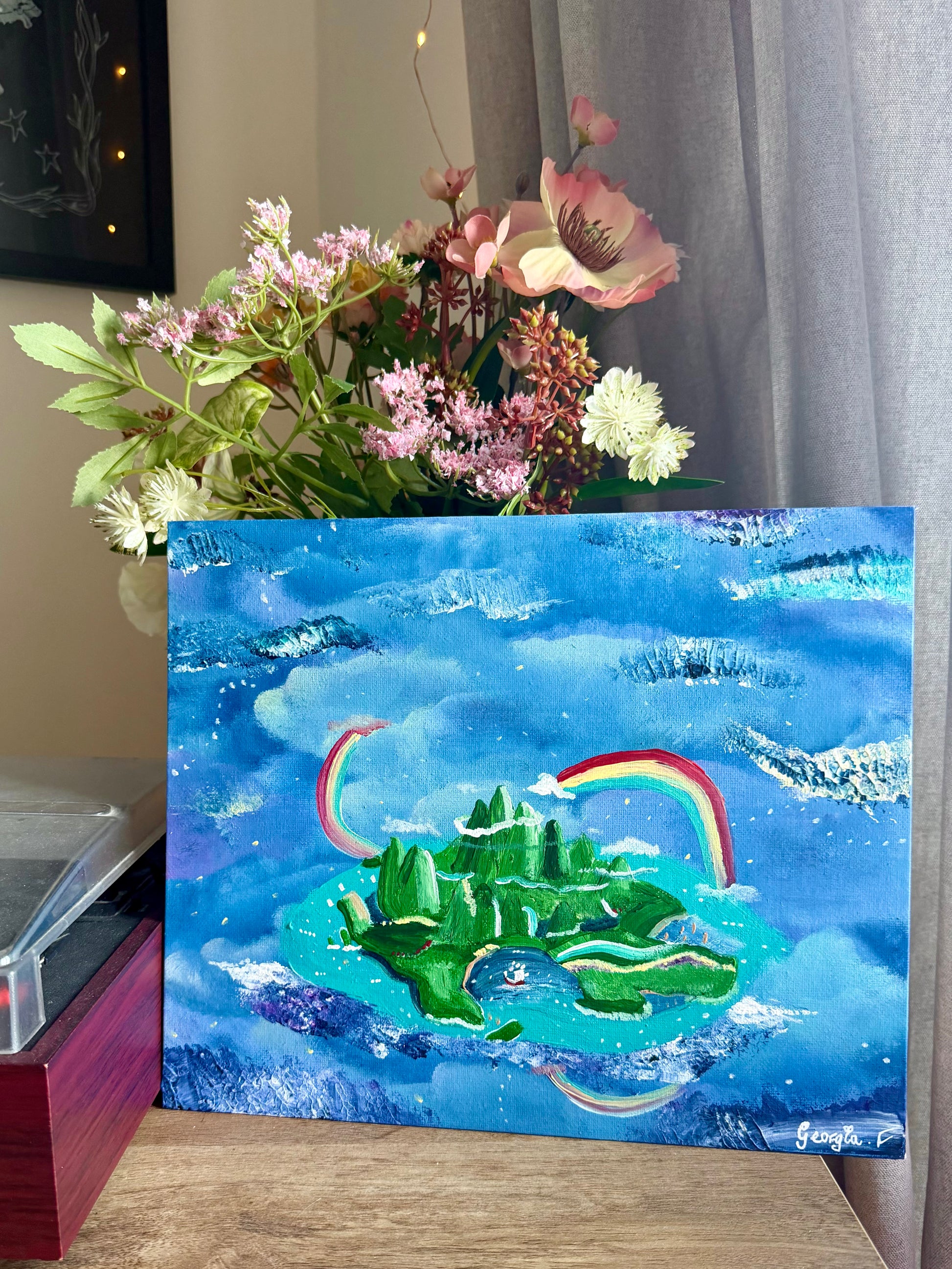 A colorful painting of a green island under a blue sky with rainbows, displayed on a wooden surface next to a vase of flowers.
