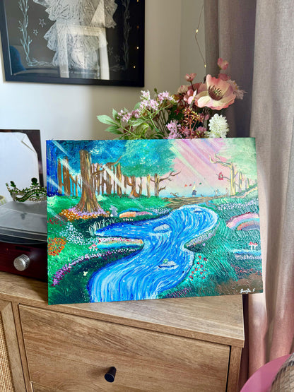 A colorful painted landscape artwork showing a winding blue river through a green forest with pink sky, displayed on a wooden cabinet in a real indoor setting with flowers and home decor visible around it.