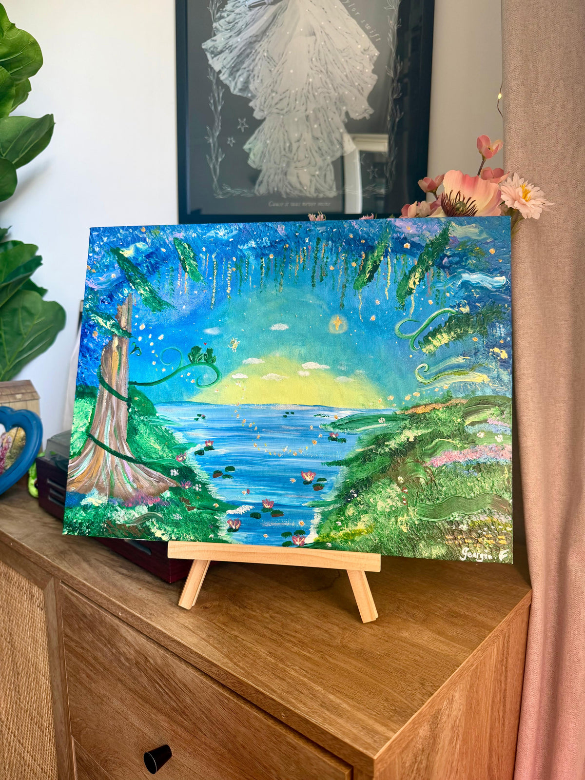 A colorful fantasy landscape painting on canvas displayed on a small wooden easel. The artwork depicts a vibrant blue and green seascape with a tree, hills, and a glowing yellow horizon.