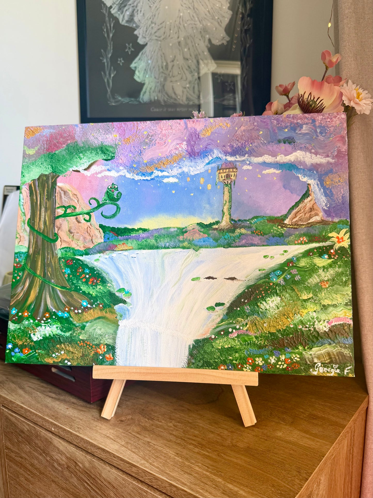 A colorful landscape painting on canvas displayed on a small wooden easel. The artwork depicts a waterfall, a large tree, flowers, hills, and a tower under a purple and blue sky.